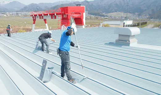 屋外で作業員が金属屋根にローラーを使って塗装を行っている様子。背景には山と施設が見える。